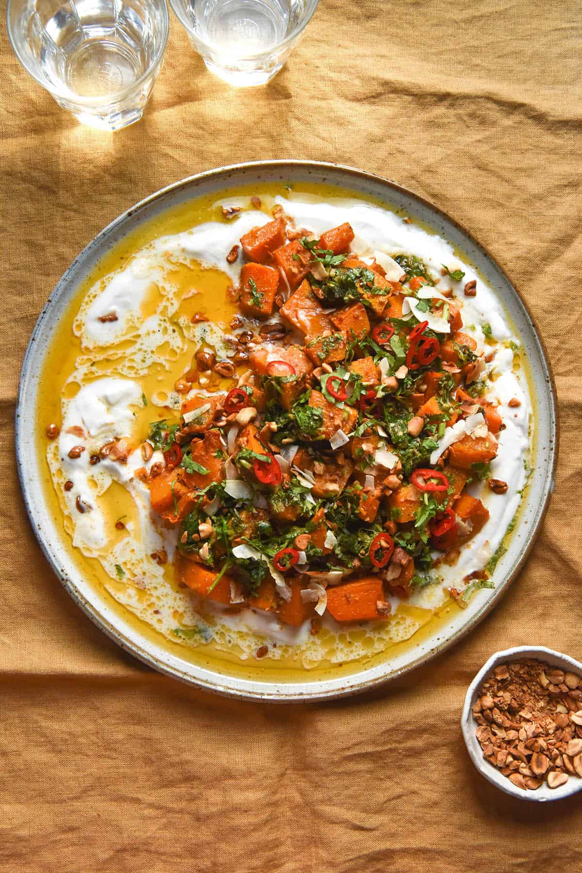 An aerial image of a white ceramic plate topped with Thai red curry roasted pumpkin on a bed of yoghurt, topped with peanuts, herbs, chilli and a lime coriander salsa. The plate sits atop a turmeric coloured linen tablecloth. A small white bowl of peanuts sits to the bottom right of the image, while two sunlit water glasses sit in the top of the image.