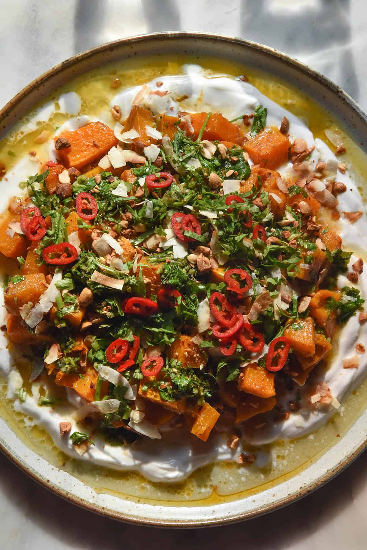 An aerial close up image of a a white ceramic plate topped with Thai red curry roasted pumpkin on a bed of yoghurt, topped with peanuts, herbs, chilli and a lime coriander salsa. The plate sits atop a white marble table.