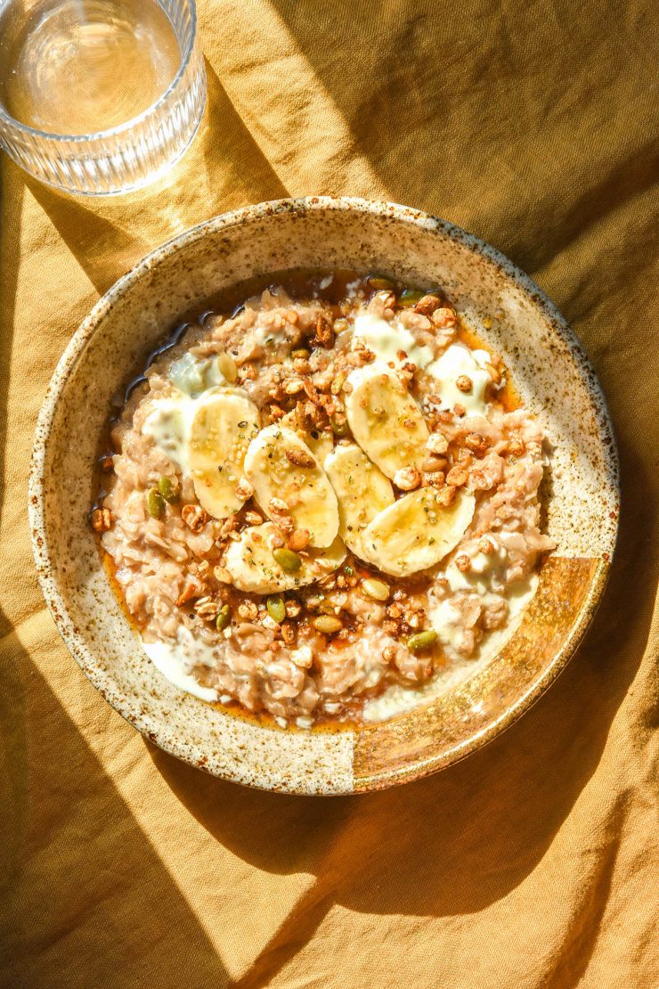 An aerial image of a bowl of vegan gluten free porridge on a mustard coloured tablecloth in bright sunlight. The porridge is topped with banana, yoghurt, seeds and maple syrup.
