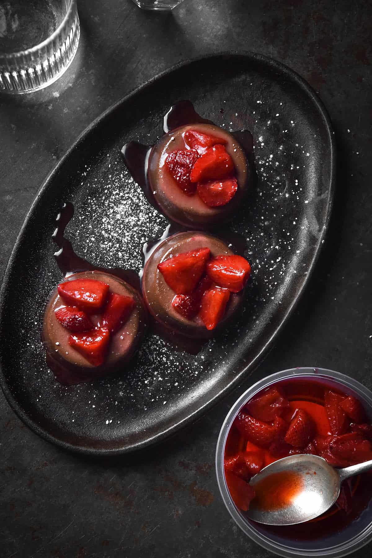 An aerial image of three chocolate panna cotta on a dark steel plate atop a dark metal backdrop. The panna cotta are topped with strawberries and a sprinkle of icing sugar. 