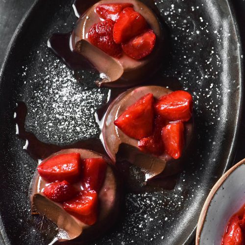 An aerial image of three chocolate panna cotta on a dark steel plate atop a dark metal backdrop. The panna cotta are topped with strawberries and a sprinkle of icing sugar. A spoonful of each panna cotta has been taken, revealing the creamy innards.