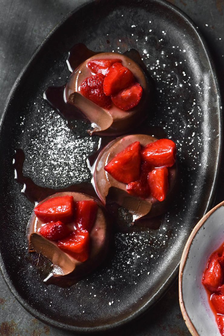 An aerial image of three chocolate panna cotta on a dark steel plate atop a dark metal backdrop. The panna cotta are topped with strawberries and a sprinkle of icing sugar. A spoonful of each panna cotta has been taken, revealing the creamy innards.