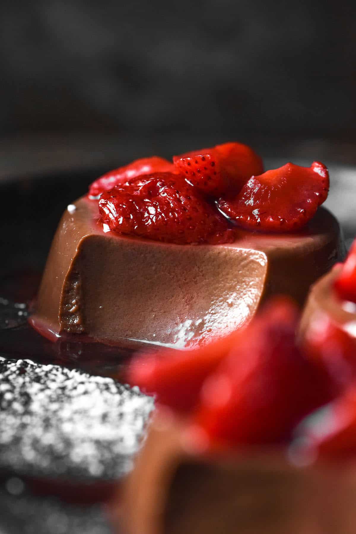 A side on image of chocolate panna cottas on a dark steel plate. The panna cottas are topped with cooked strawberries and icing sugar. A spoonful of each panna cotta has been taken, revealing the creamy innards.