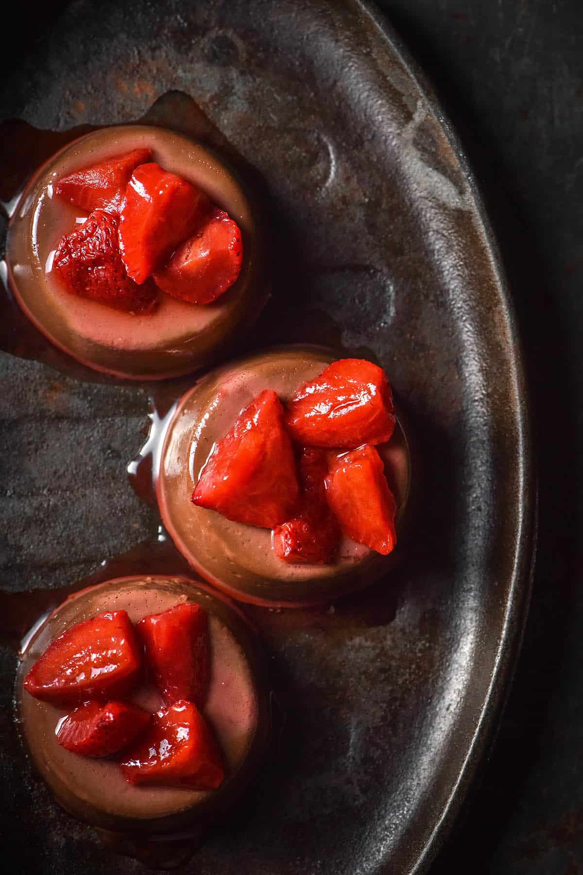 An aerial image of three chocolate panna cotta on a dark steel plate atop a dark backdrop. The panna cotta are topped with cooked strawberries,