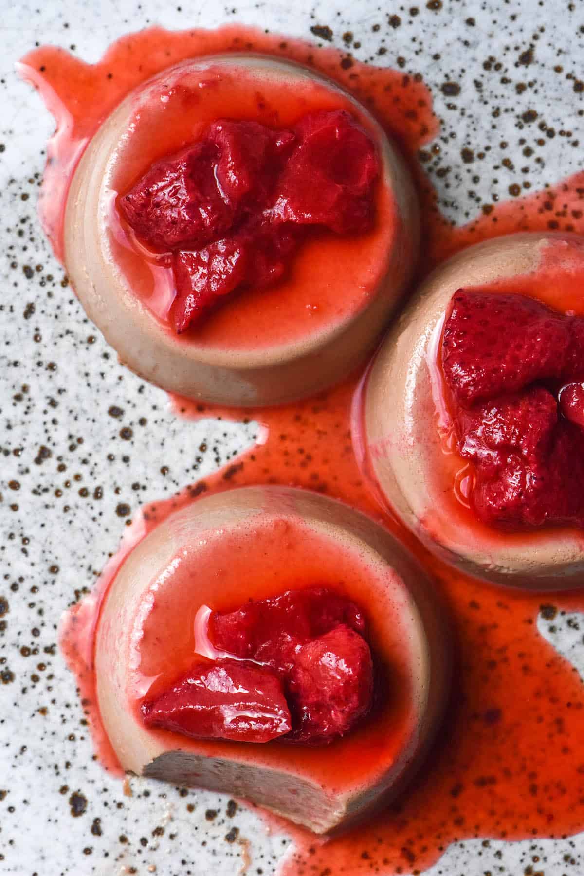 An aerial image of three chocolate panna cotta on a white speckled ceramic plate. The panna cotta are topped with cooked strawberries.