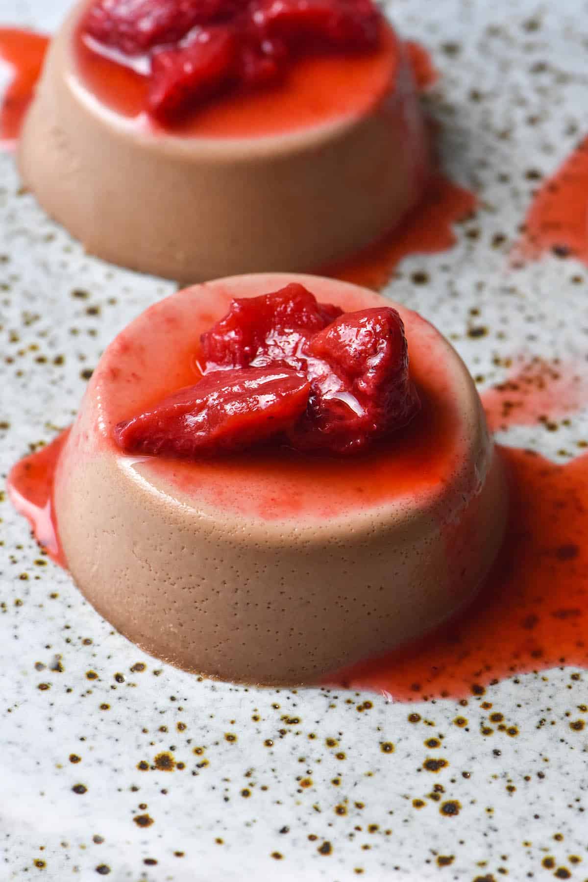 An aerial side on image of two chocolate panna cotta on a white speckled ceramic plate. The panna cotta are topped with cooked strawberries.