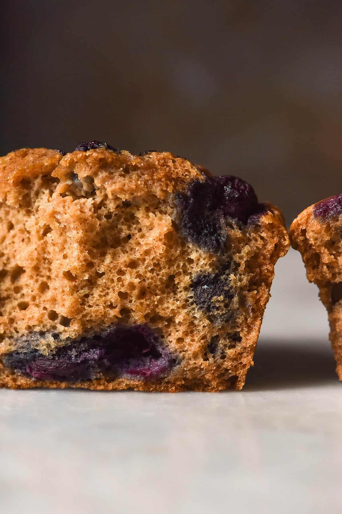 A side on image of a buckwheat blueberry muffin that has been torn in half to reveal the soft inner crumb studded with blueberries. The muffin halves sit atop a white marble table against a dark backdrop.