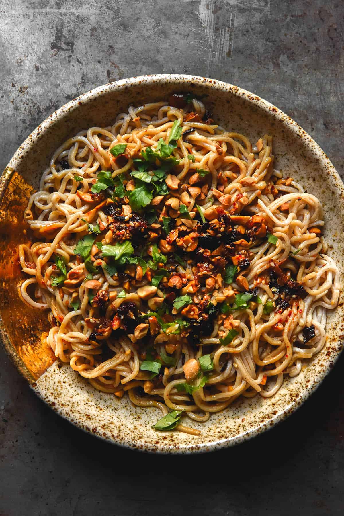 An aerial image of gluten free egg noodles in a beige speckled ceramic bowl. The egg noodles are topped with chilli crisp. peanuts and chopped coriander and the bowl sits atop a dark steel backdrop
