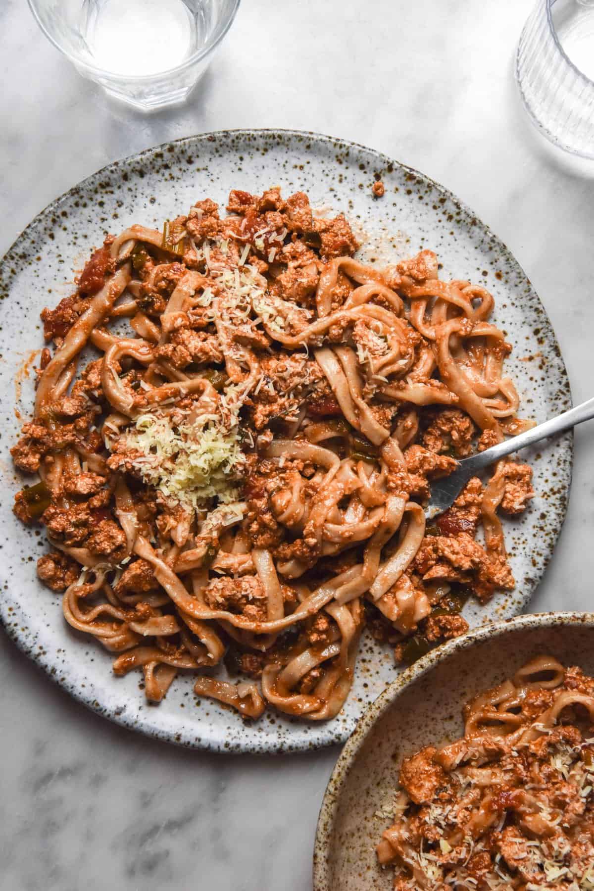 An aerial image of gluten free egg pasta topped with vegetarian bolognese and parmesan on a white speckled ceramic plate atop a white marble table. The plate is surrounded by an extra bowl of bolognese and glasses of water