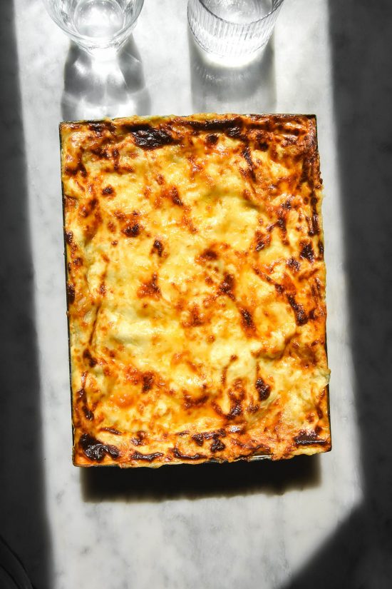 A sunlit aerial image of a gluten free lasagne made with gluten free lasagne sheets. The lasagne sits on a white marble table in contrasting sunlight and two glasses of water sit to the top of the image