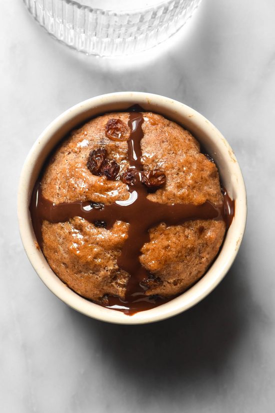 An aerial image of a gluten free microwave hot cross bun with a dark chocolate cross. The bun sits on a white marble table and a sunlit glass of water sits to the top of the image.