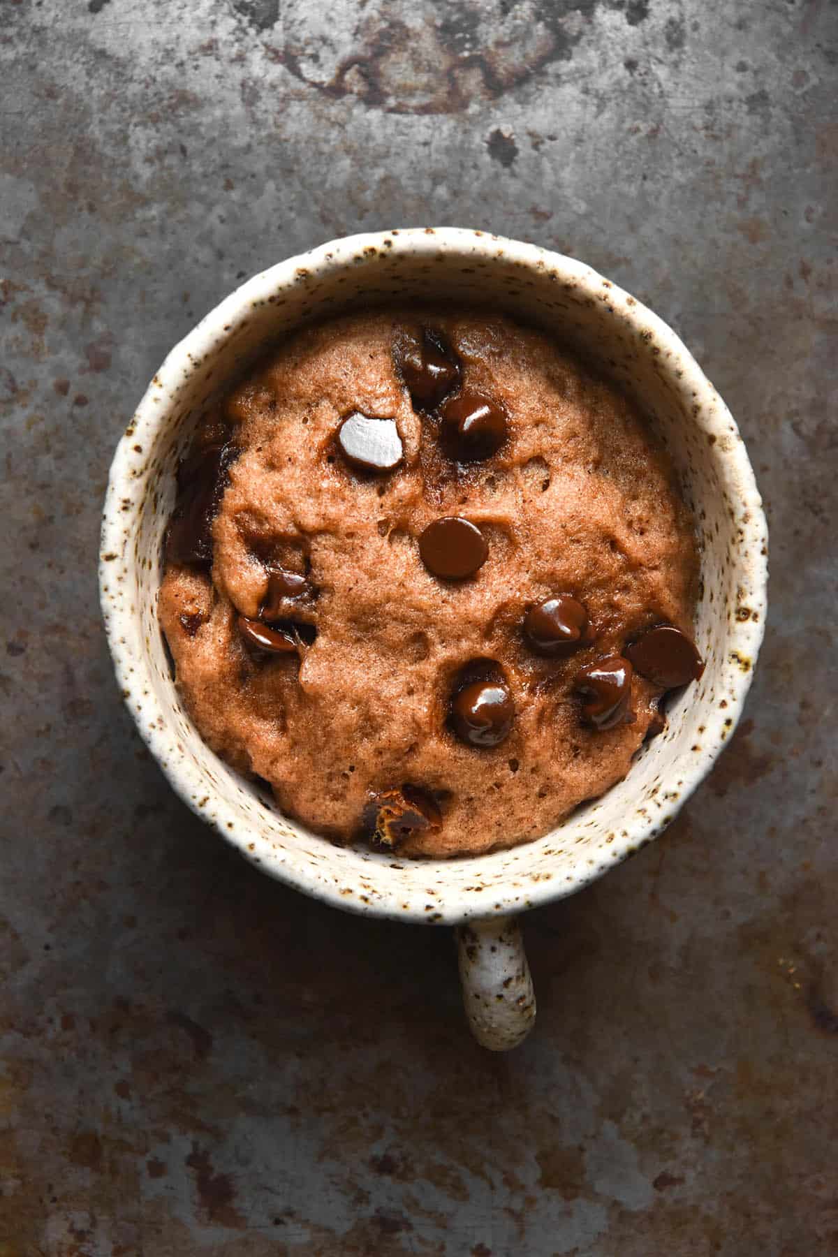 An aerial image of a gluten free microwave hot cross bun topped with chocolate chips. The bun sits in a white speckled ceramic mug atop a dark steel backdrop