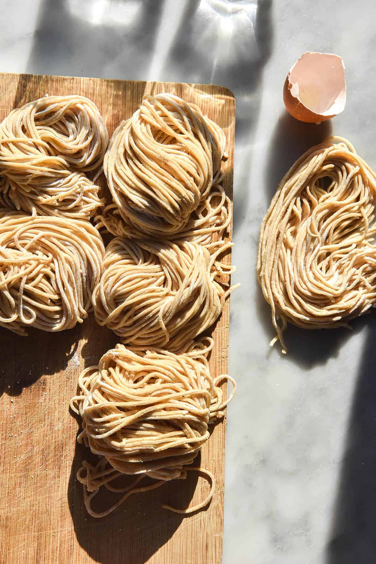 An aerial image of gluten free noodles on a wooden board atop a white marble table in contrasting sunlight