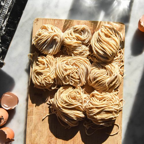 An aerial sunlit image of nests of gluten free egg noodles on a wooden chopping board atop a white marble table in bright sunlight. The noodles are surrounded by eggs, glasses of water and a pasta machine