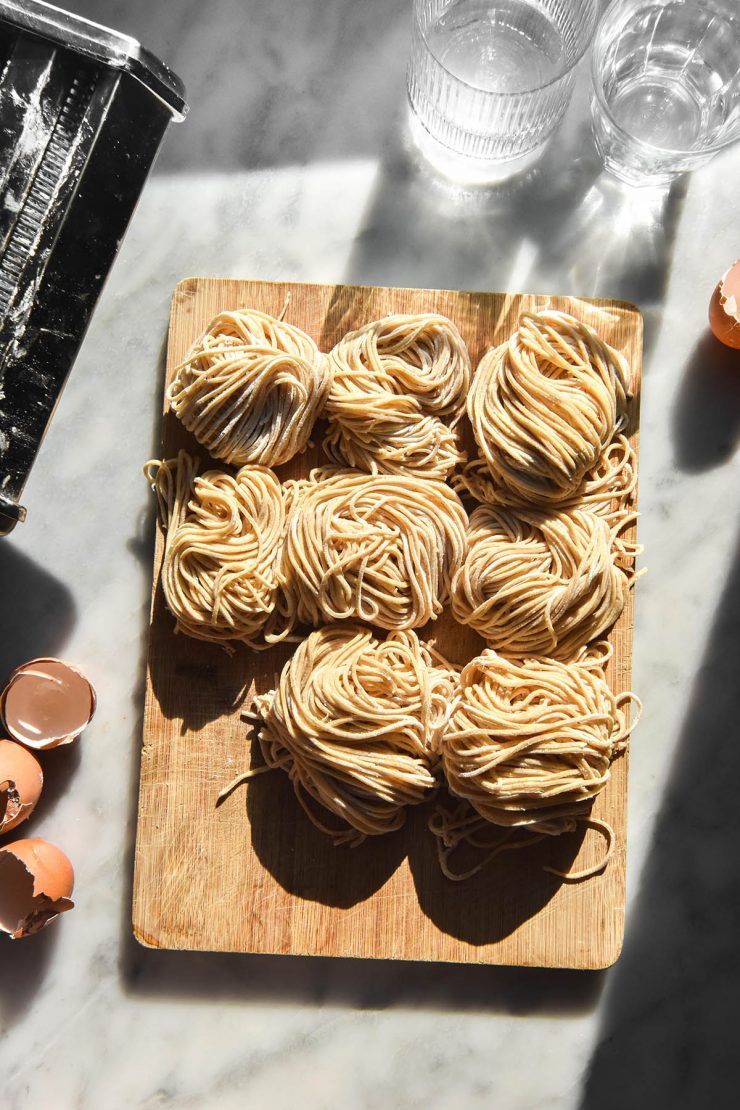 An aerial sunlit image of nests of gluten free egg noodles on a wooden chopping board atop a white marble table in bright sunlight. The noodles are surrounded by eggs, glasses of water and a pasta machine