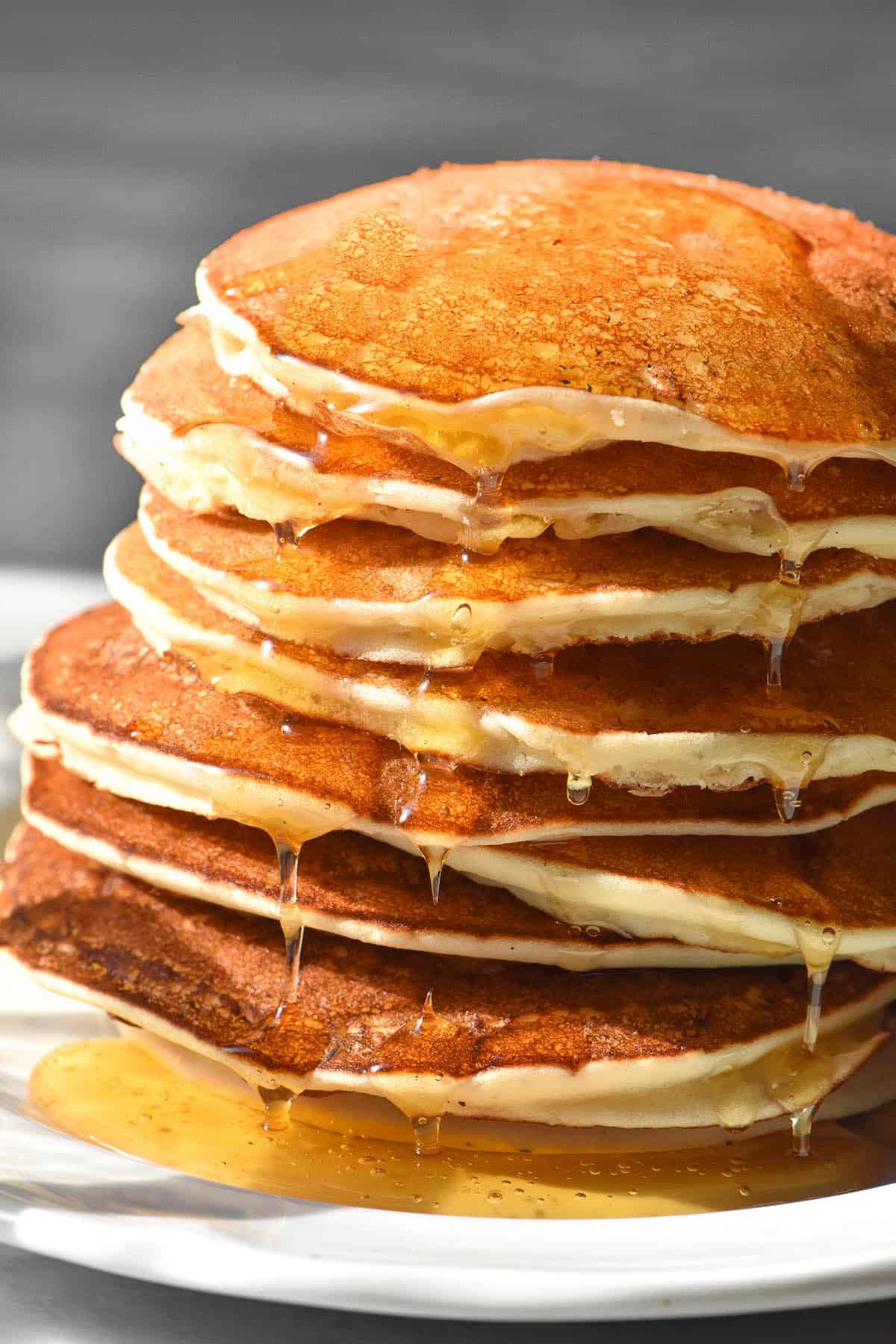 A side on image of a stack of gluten free ricotta pancakes on a white plate atop a white marble table. The pancakes have been drizzled with honey which drips down the sides of the stack.
