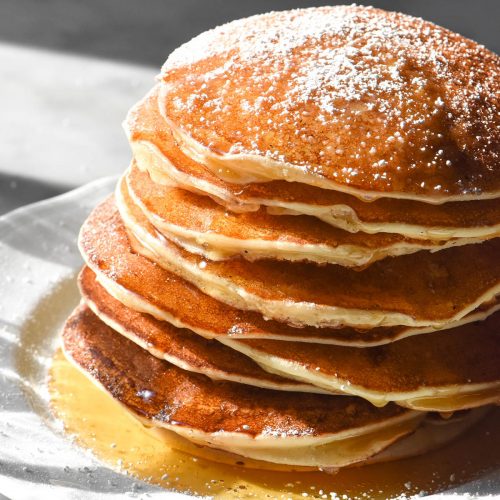 A side on image of gluten free ricotta pancakes drizzled with honey and sprinkled with icing sugar. The stack sits on a white plate atop a white marble table in contrasting sunlight.
