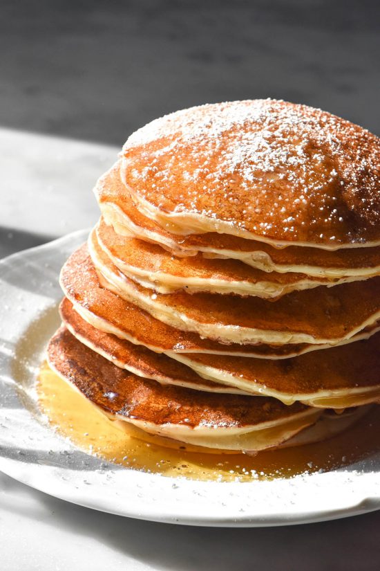 A side on image of gluten free ricotta pancakes drizzled with honey and sprinkled with icing sugar. The stack sits on a white plate atop a white marble table in contrasting sunlight.