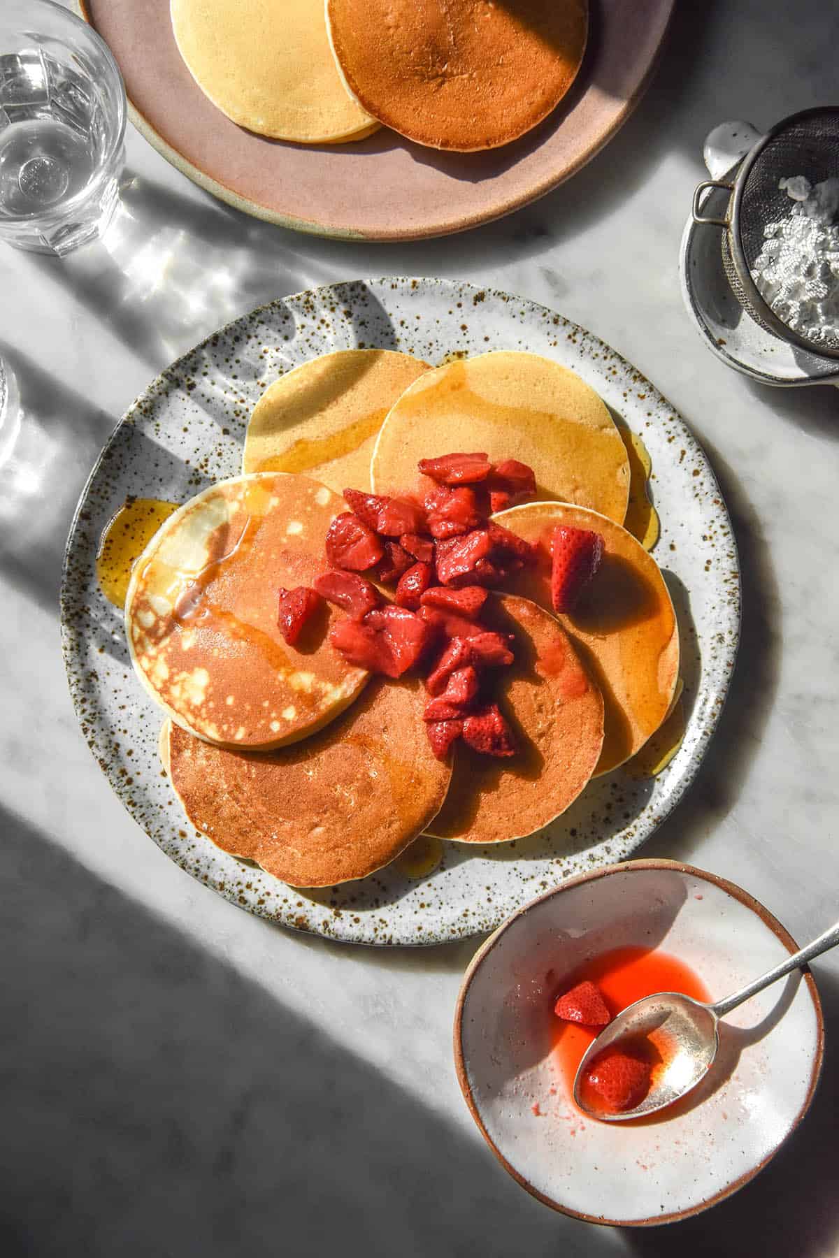 An aerial image of a white speckled ceramic plate on a white marble table topped with gluten free ricotta pancakes. The pancakes are topped with stewed strawberries.