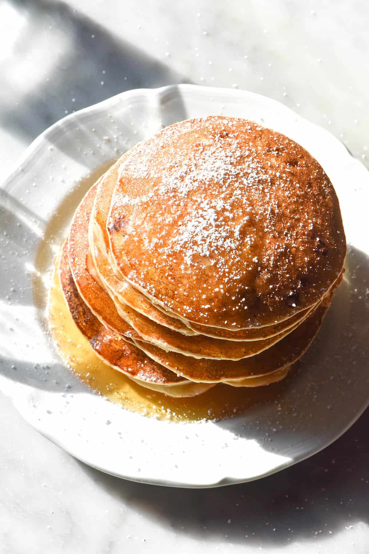 An aerial image of gluten free ricotta pancakes drizzled with honey and sprinkled with icing sugar. The stack sits on a white plate atop a white marble table in contrasting sunlight.