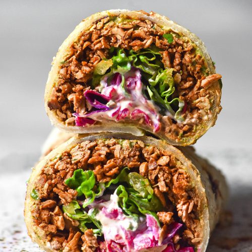A side on image of a gluten free tortilla filled with tofu mince, pickled red cabbage, lettuce and sour cream. The burrito has been sliced in half, revealing the insides. The burrito pieces are stacked on top of one another on a white plate atop a white marble table