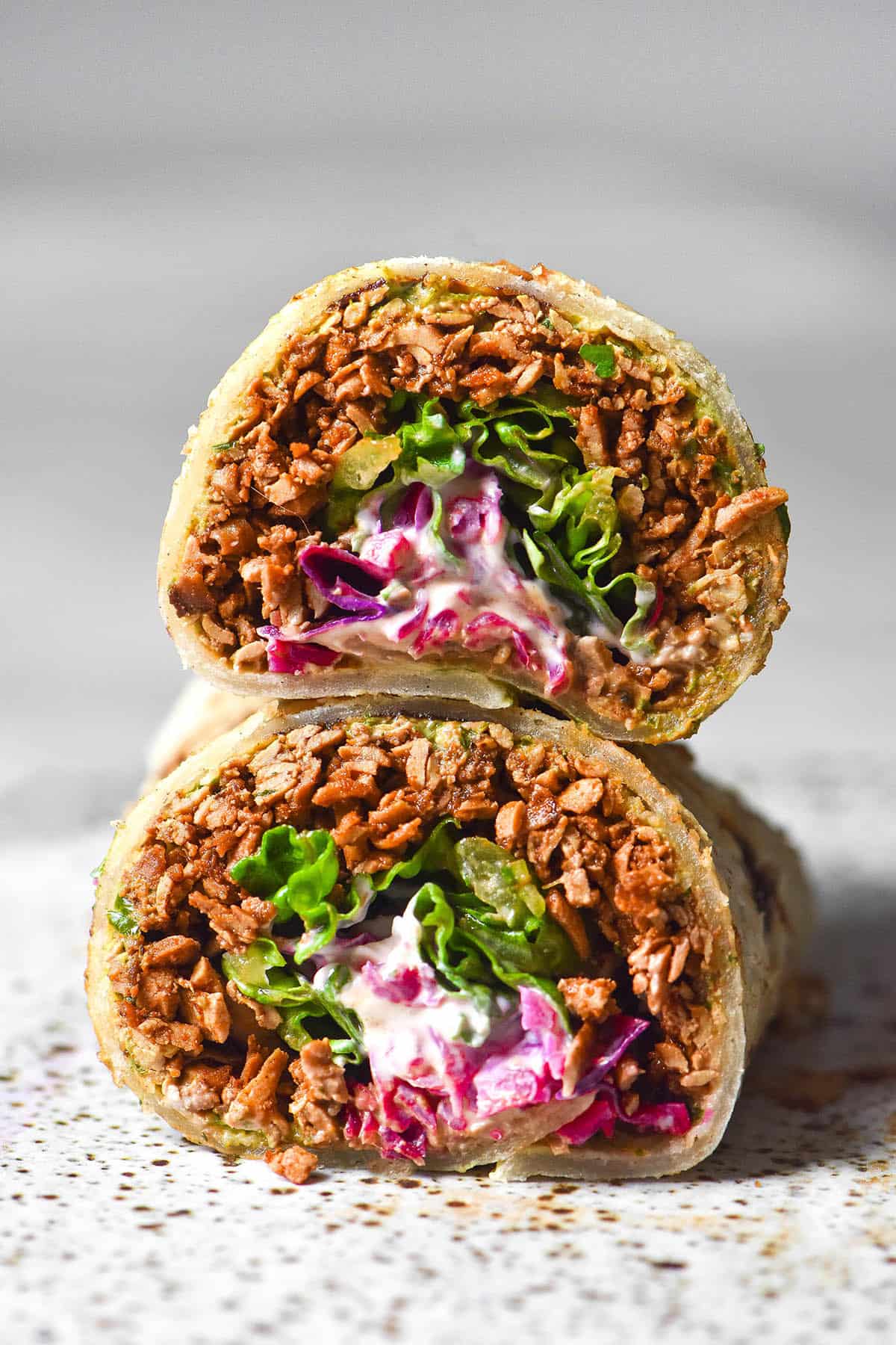 A side on image of a gluten free tortilla filled with tofu mince, pickled red cabbage, lettuce and sour cream. The burrito has been sliced in half, revealing the insides. The burrito pieces are stacked on top of one another on a white plate atop a white marble table