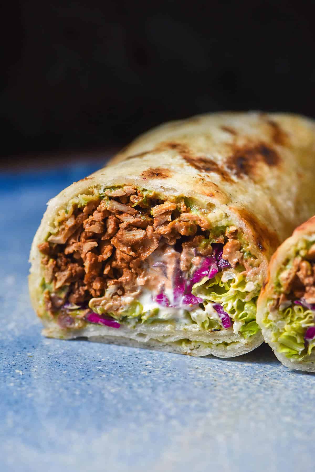 A side on image of half a gluten free burrito stuffed with tofu mince, pickled cabbage, lettuce, an avocado cilantro dressing and sour cream. The burrito sits on a bright blue ceramic plate against a dark backdrop