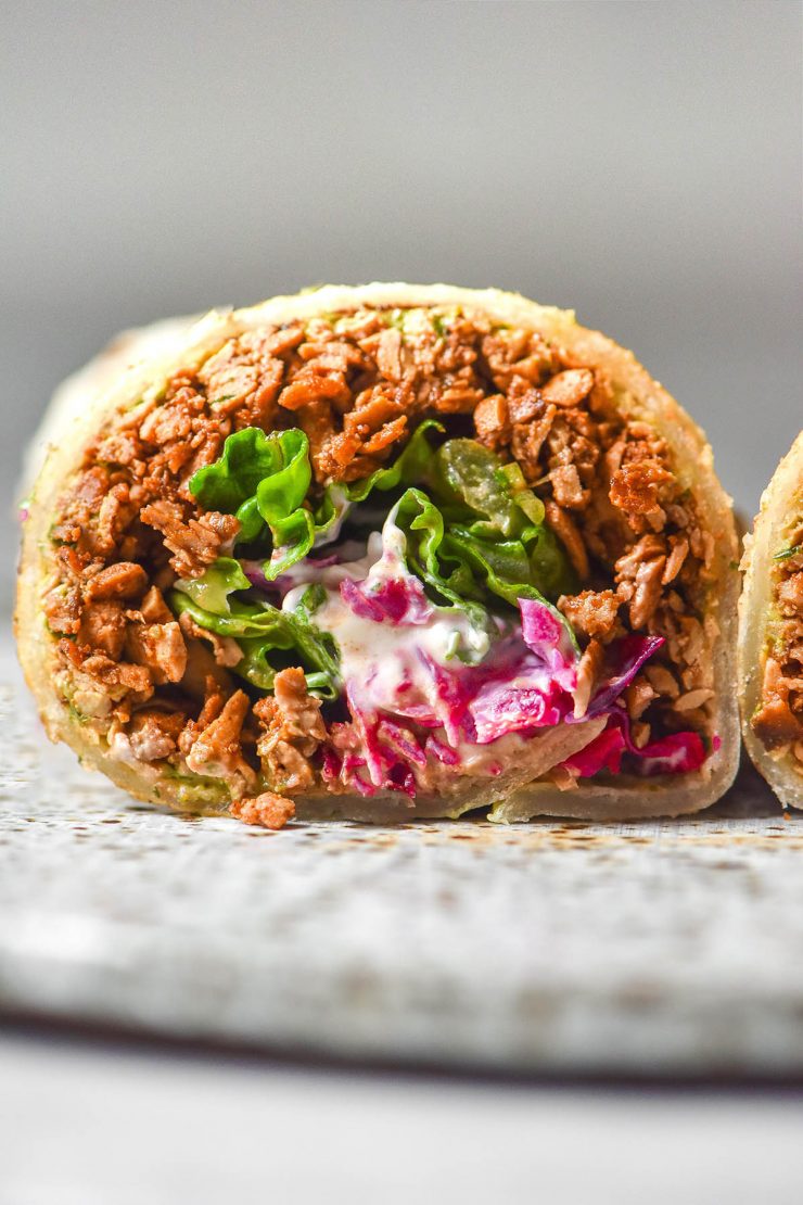 A side on image of a gluten free tortilla filled with tofu mince, pickled red cabbage, lettuce and sour cream. The burrito has been sliced and sits on a white plate atop a white marble table