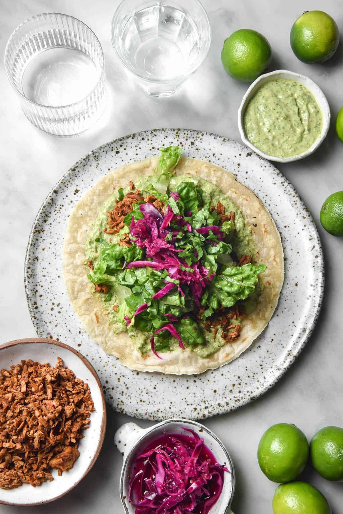 An aerial image of a gluten free tortilla topped with tofu mince, lettuce and pickled red cabbage. The tortilla sits atop a white speckled ceramic plate atop a white marble table. The plate is surrounded by bowls of the ingredients, extra limes and glasses of water.