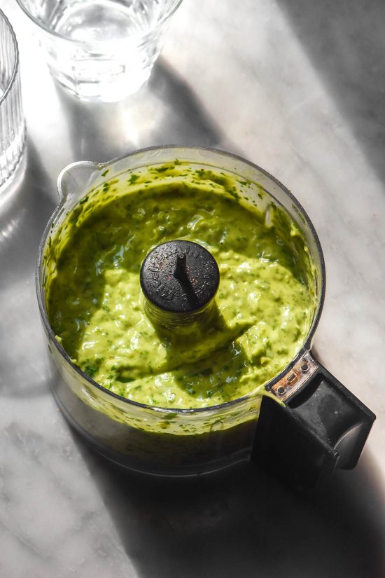 An aerial sunlit image of low FODMAP avocado cilantro dressing in a food processor on a white marble table. Two glasses of water sit to the top left of the image, creating contrasting light and shadow.
