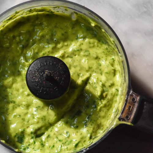 An aerial sunlit image of low FODMAP avocado cilantro dressing in a food processor on a white marble table