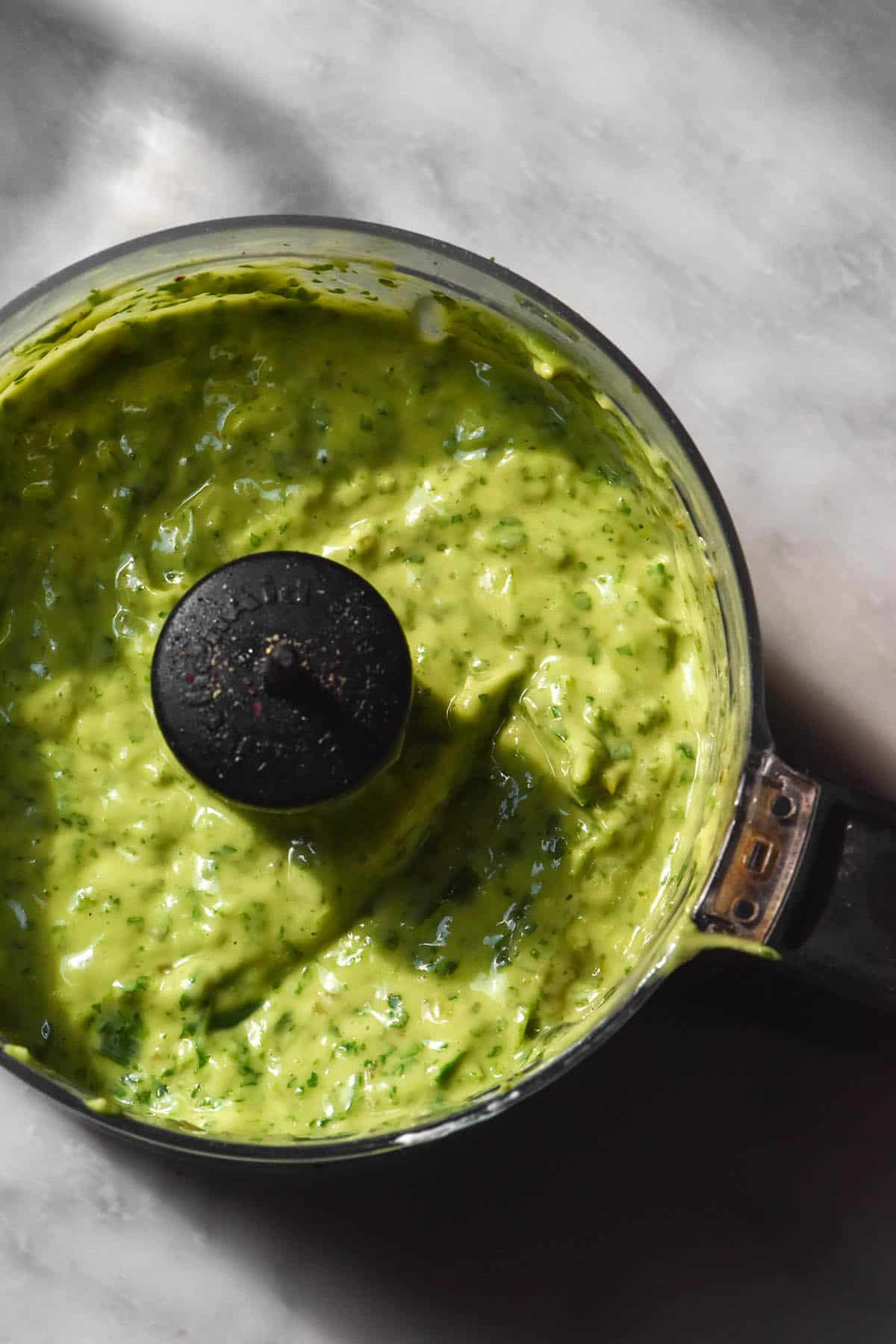 An aerial sunlit image of low FODMAP avocado cilantro dressing in a food processor on a white marble table