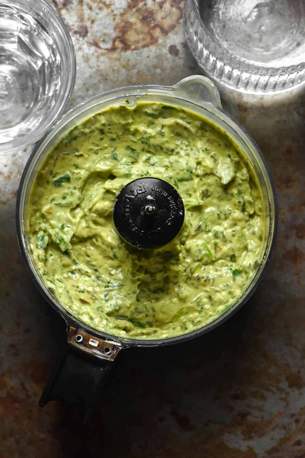 An aerial image of avocado cilantro sauce in a food processor on a dark steel backdrop surrounded by two glasses of water.