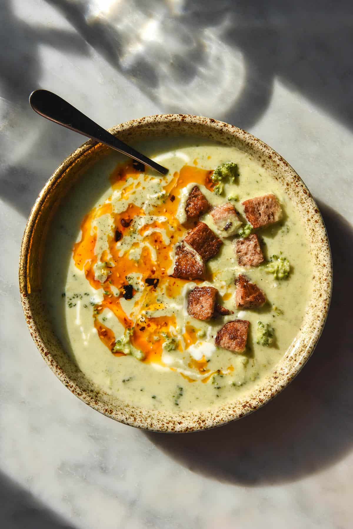 An aerial image of a beige speckled ceramic bowl on a white marble table filled with low FODMAP broccoli cheddar soup. The soup is topped with gluten free croutons, cream and low FODMAP chilli crisp