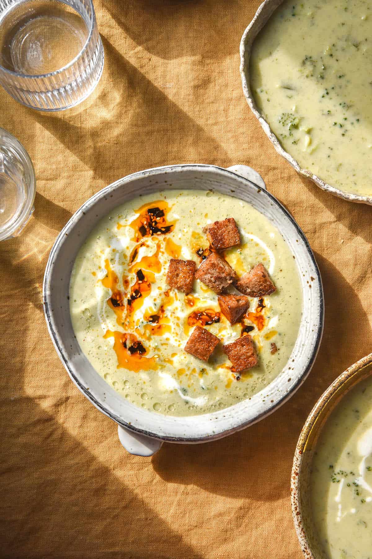A brightly lit aerial image of broccoli cheddar soup in a white bowl atop a mustard coloured linen tablecloth. The central bowl is topped with croutons and chilli oil and surrounded by other bowls of soup and sunlit water glasses.