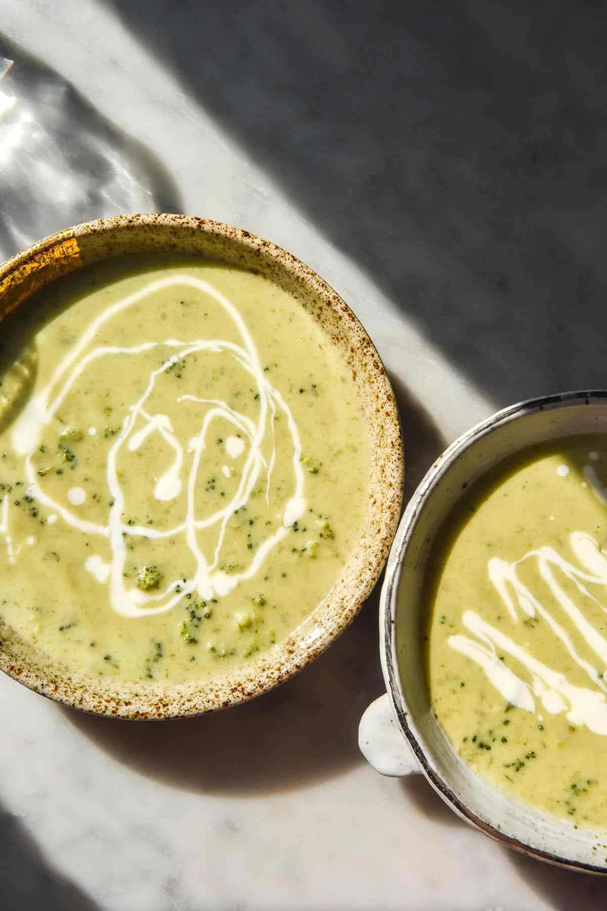 A sunlit aerial image of two bowls of low FODMAP broccoli cheddar soup on a white marble table