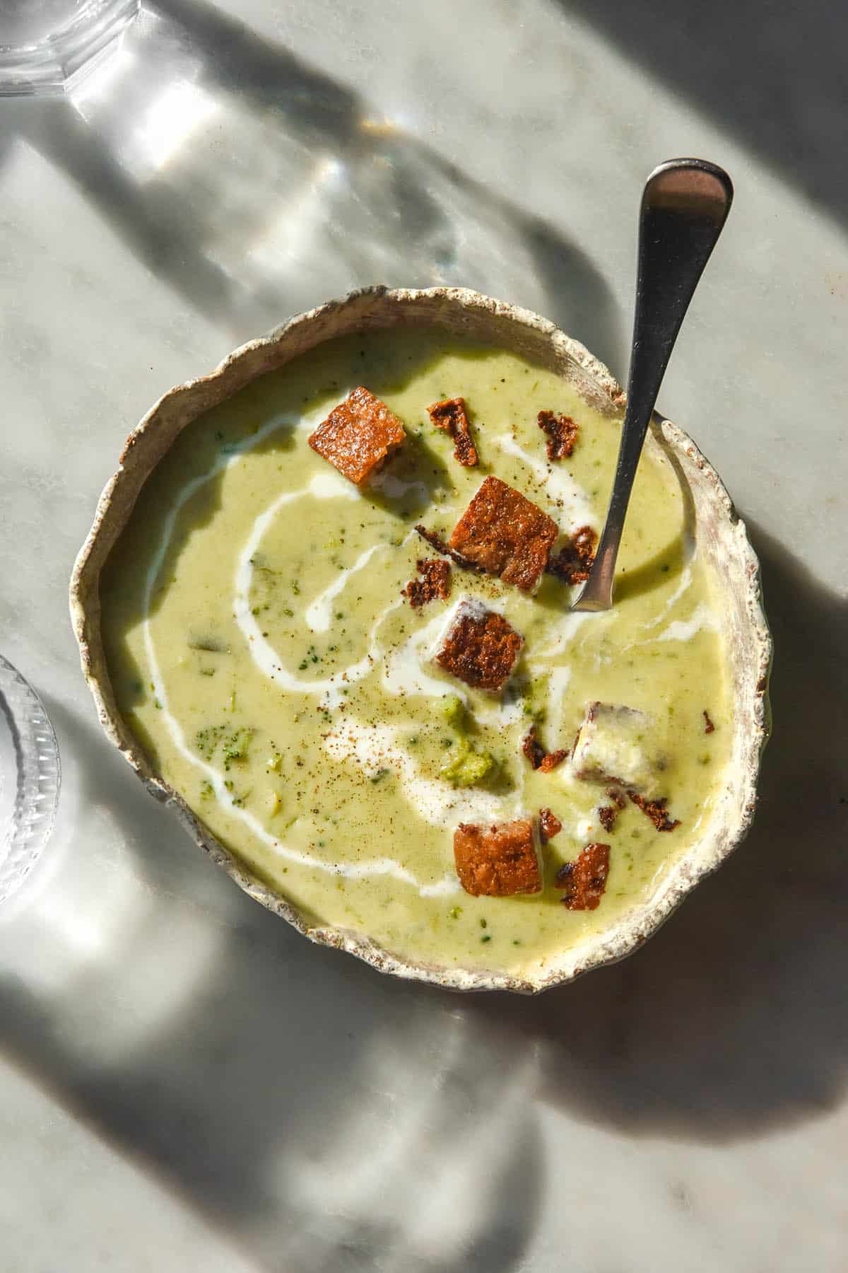 An aerial image of a white misshapen ceramic bowl filled with low FODMAP broccoli cheddar soup. The glass sits on a white marble table in contrasting sunlight.
