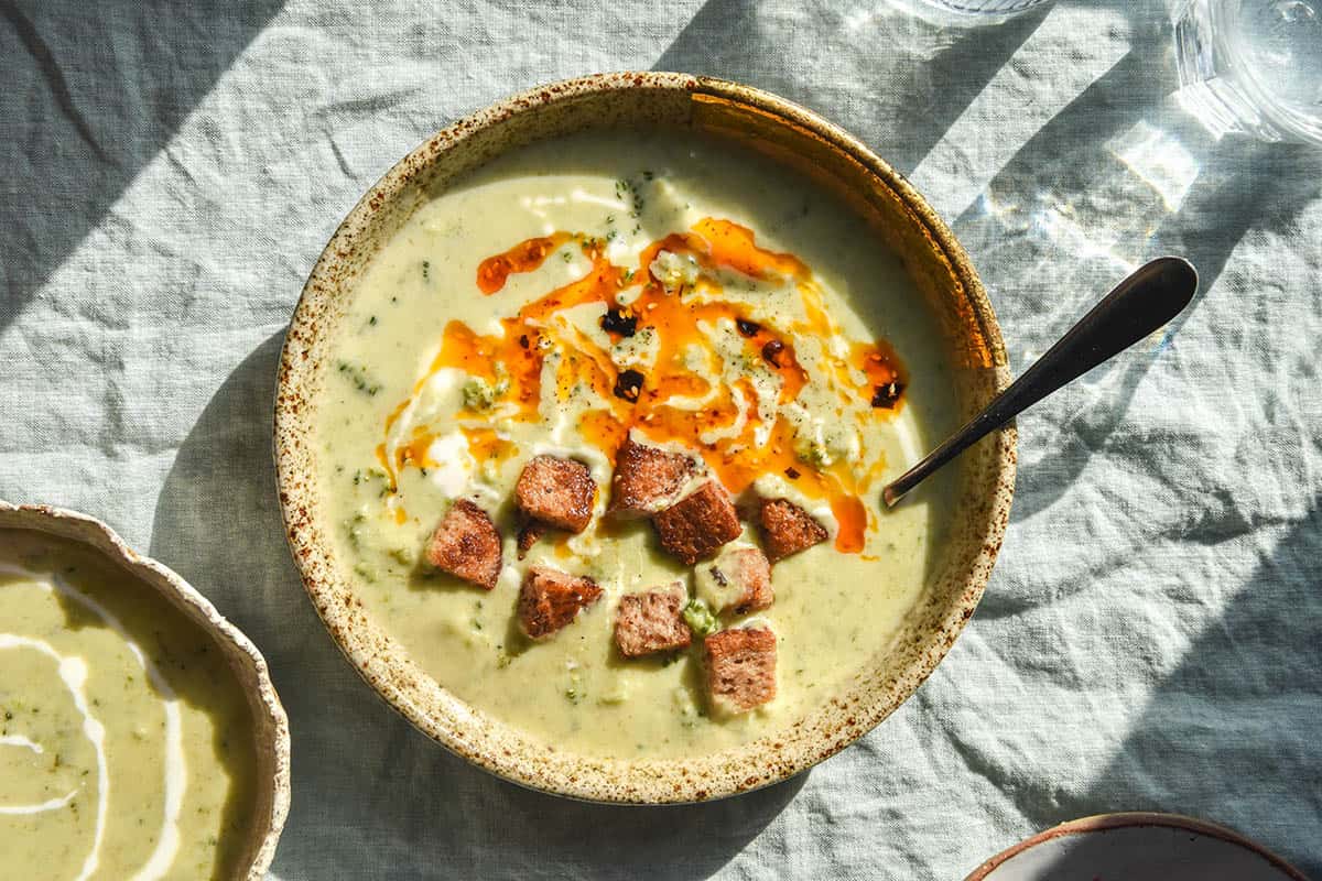 An aerial image of a beige speckled ceramic bowl on a light green linen tablecloth filled with low FODMAP broccoli cheddar soup. The soup is topped with gluten free croutons, cream and low FODMAP chilli crisp. A second bowl of soup sits to the bottom left of the image and two water glasses sit to the right.