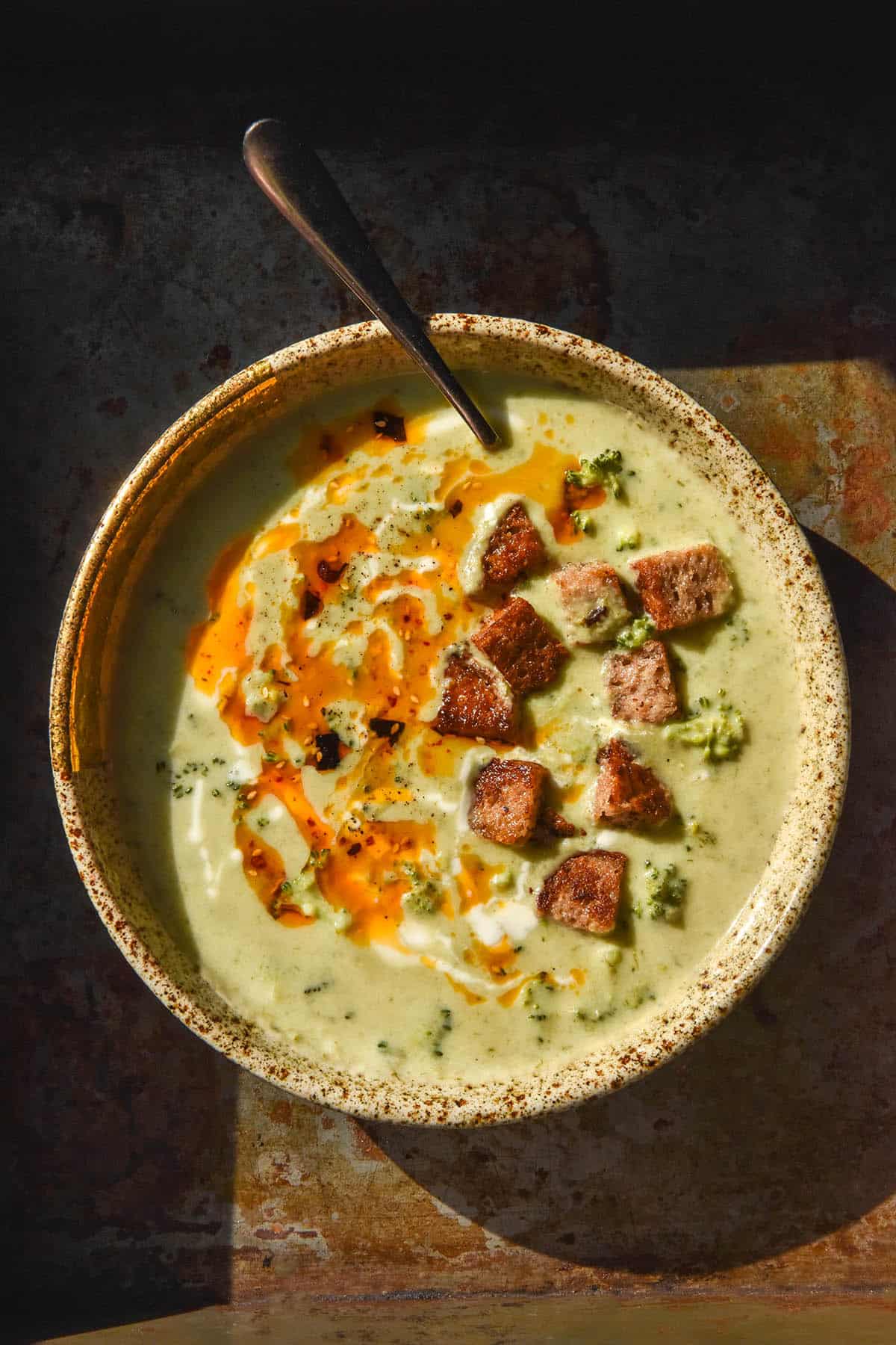An aerial image of a beige speckled ceramic bowl filled with low FODMAP broccoli cheddar soup. The soup is topped with cream, low FODMAP chilli crisp and gluten free croutons and sits on a dark steel backdrop in contrasting sunlight.