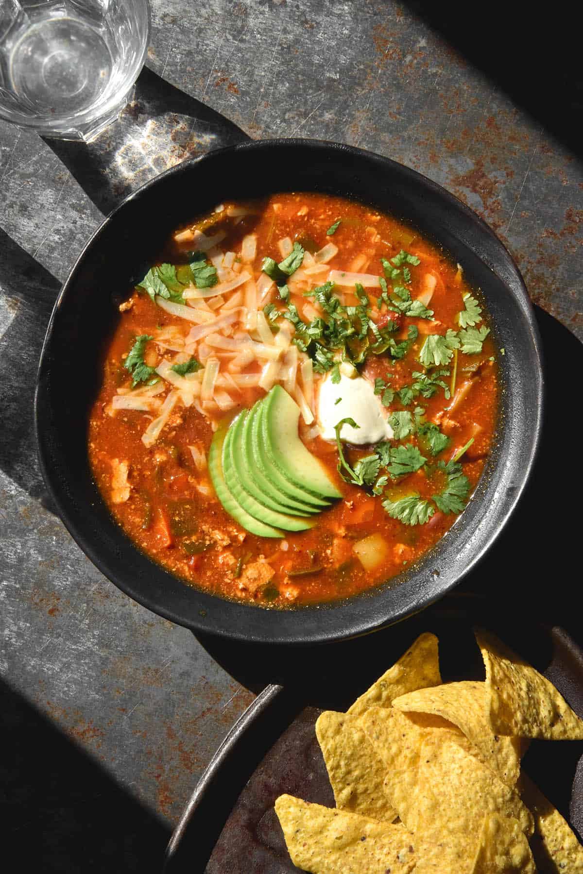 An aerial image of a dark blue ceramic bowl of vegetarian taco soup atop a dark blue backdrop. The soup is topped with grated cheese, sour cream, avocado and extra coriander. A plate of corn chips sit to the bottom right of the image.