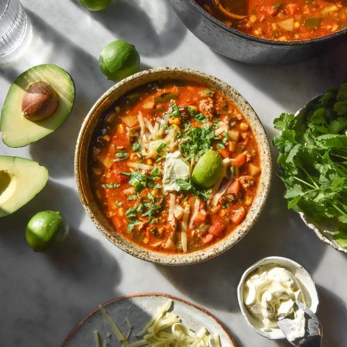 An aerial image of a bowl of low FODMAP vegetarian taco soup on a white marble table surrounded by topping ingredients, water glasses and a pot of the soup.