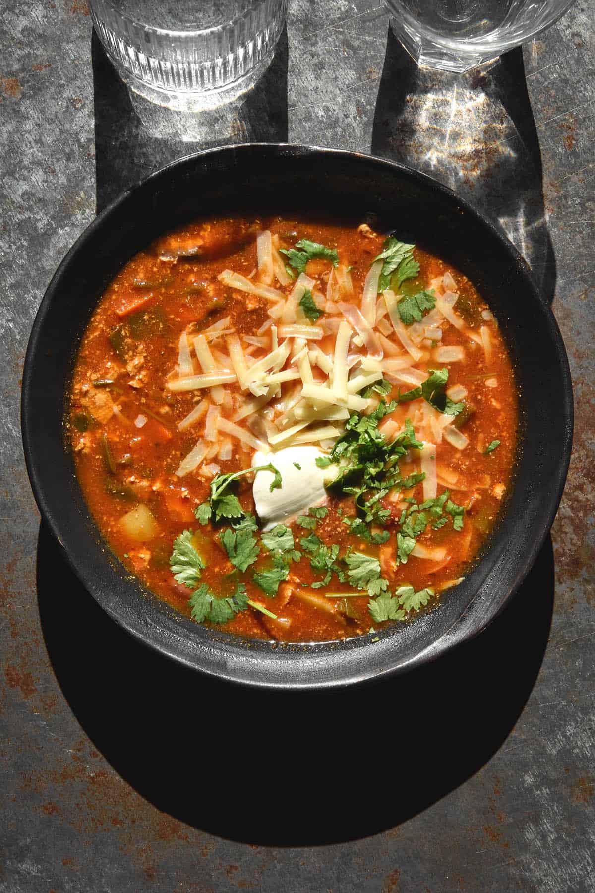 An aerial image of a dark blue ceramic bowl of vegetarian taco soup atop a dark blue backdrop. The soup is topped with grated cheese, sour cream and extra coriander.