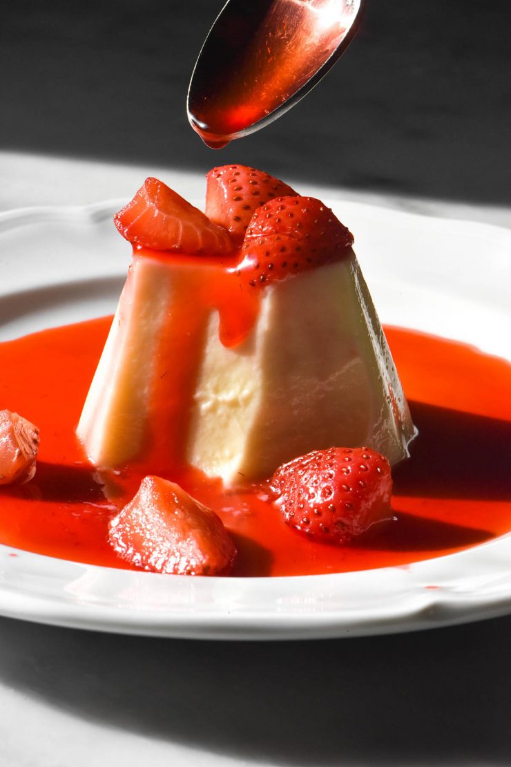 A side on image of a white chocolate panna cotta on a white plate in contrasting sunlight. The panna cotta is topped with a strawberry coulis and a spoon extends from the top of the image to drizzle more coulis over the top