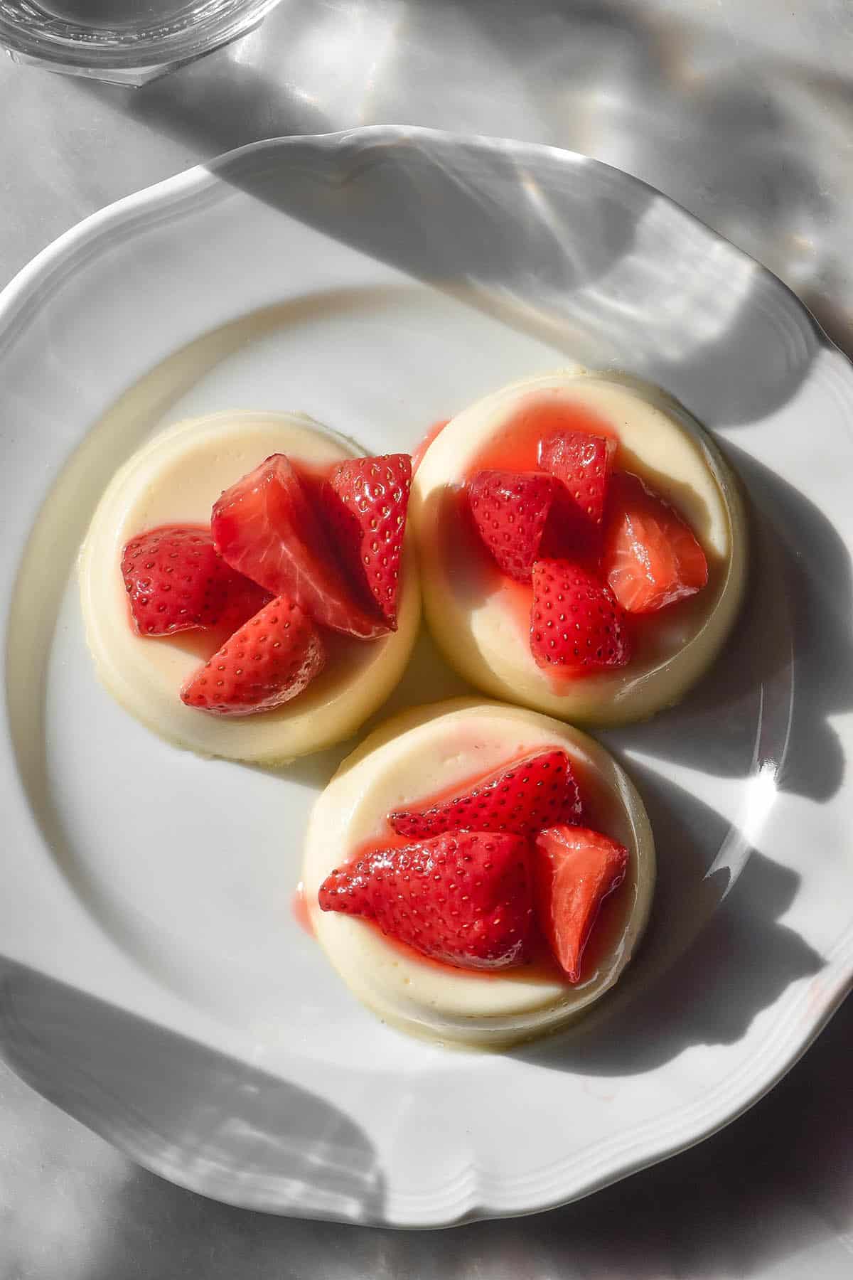 An aerial image of three white chocolate panna cotta on a white scalloped plate atop a white marble table. Sunlight shines through glasses of water at the top and bottom of the image.