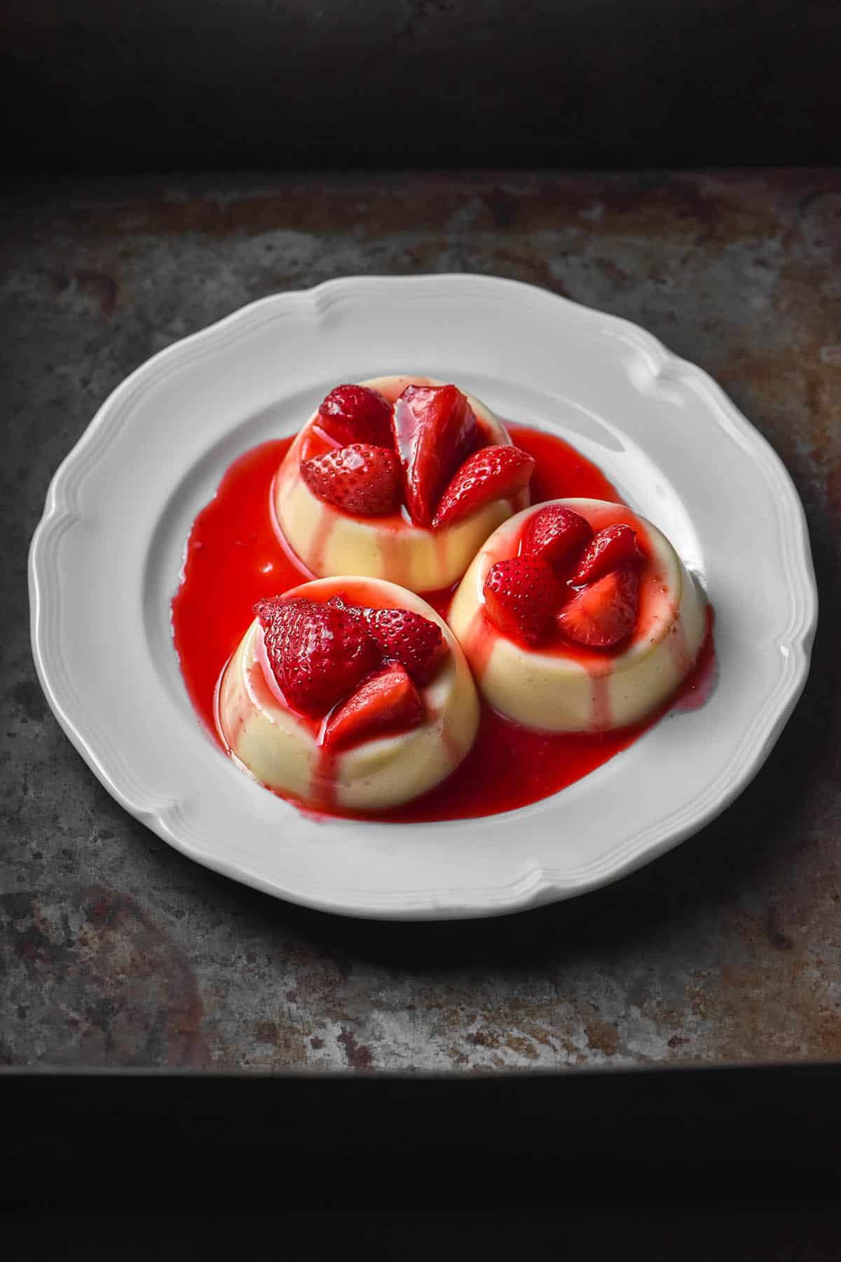 Three white chocolate panna cotta topped with strawberry coulis on a white scalloped plate atop a dark steel backdrop