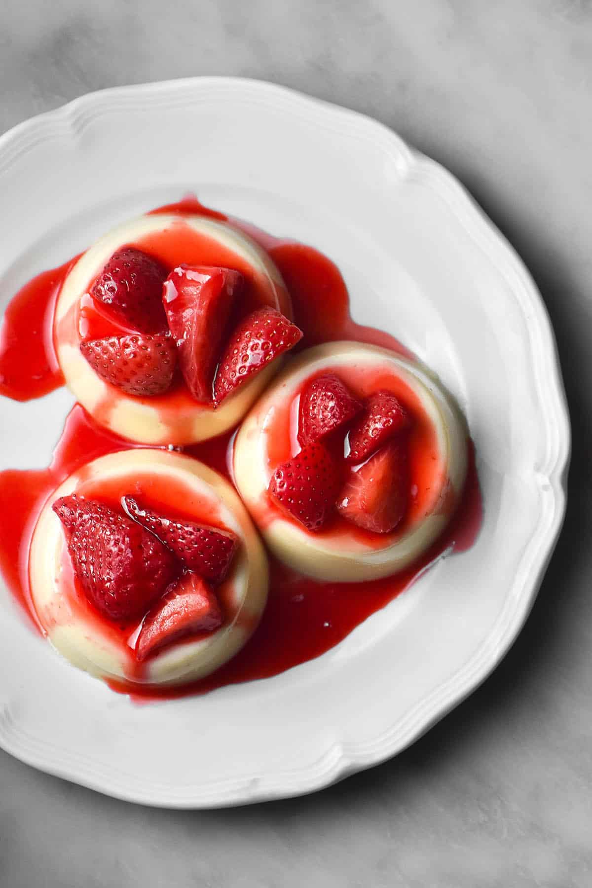 An aerial image of three white chocolate panna cotta on a scalloped white plate atop a white marble table. The panna cotta are topped with cooked strawberries with a bright red sauce