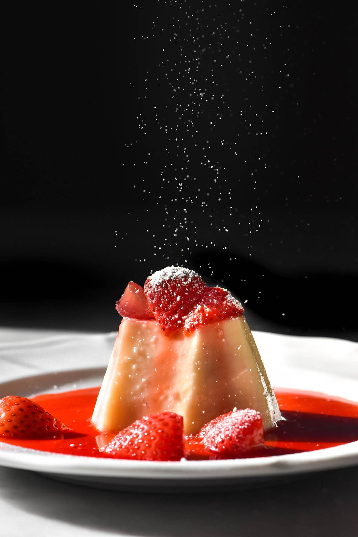 A side on image of a white chocolate panna cotta on a white plate in contrasting sunlight. The panna cotta is topped with a strawberry coulis and icing sugar that is being sprinkled from the top of the image