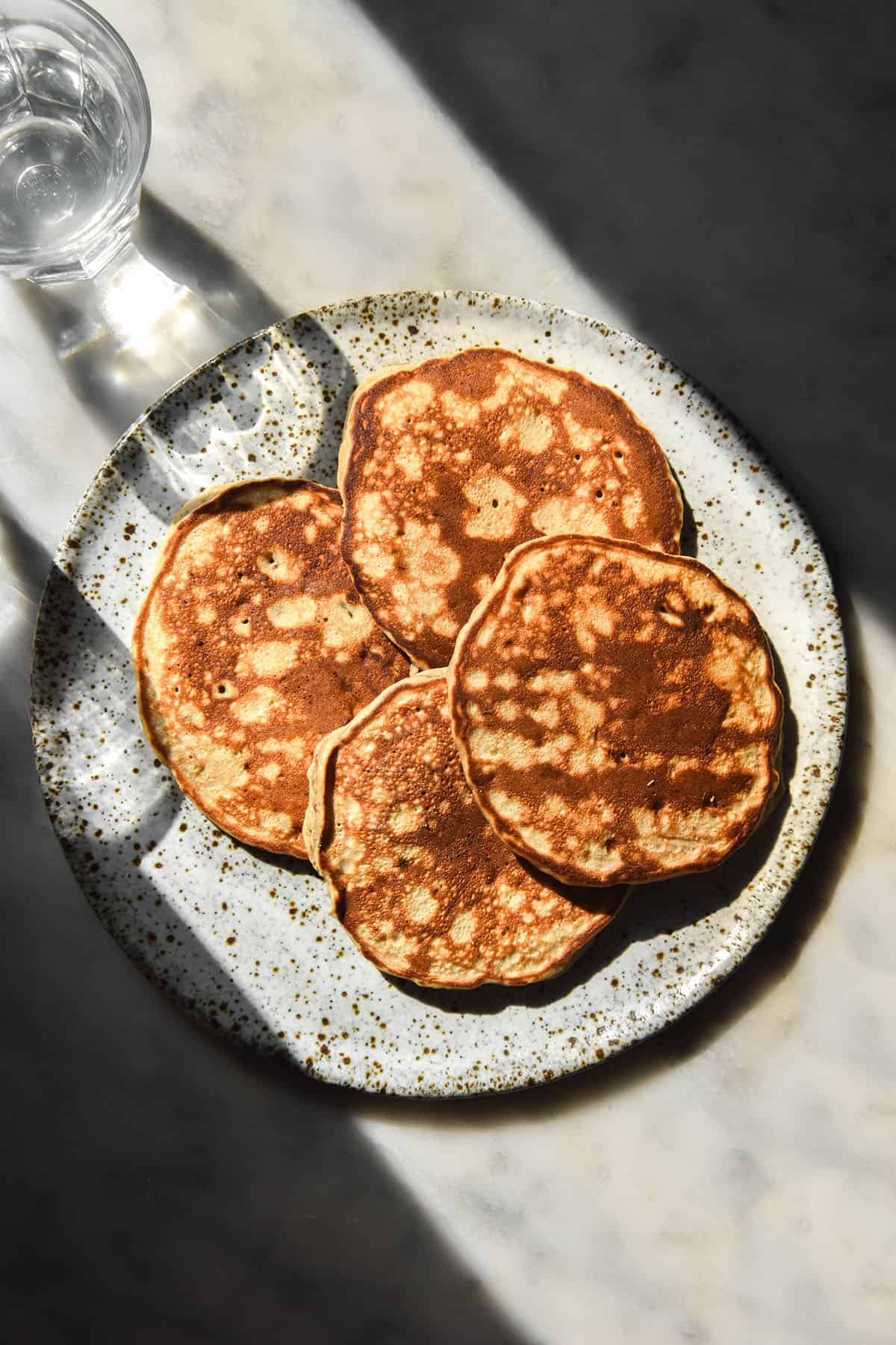 An aerial image of buckwheat banana pancakes in contrasting bright sunlight. The pancakes sit on a white speckled ceramic plate framed by water glasses and dark shadows to either side of the plate.