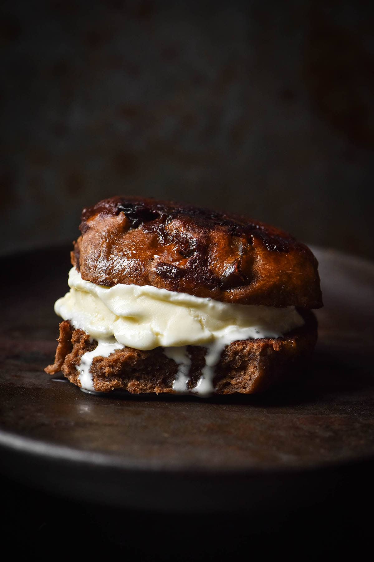 A moody side on image of a gluten free hot cross bun toasted and sandwiched with vanilla ice cream.