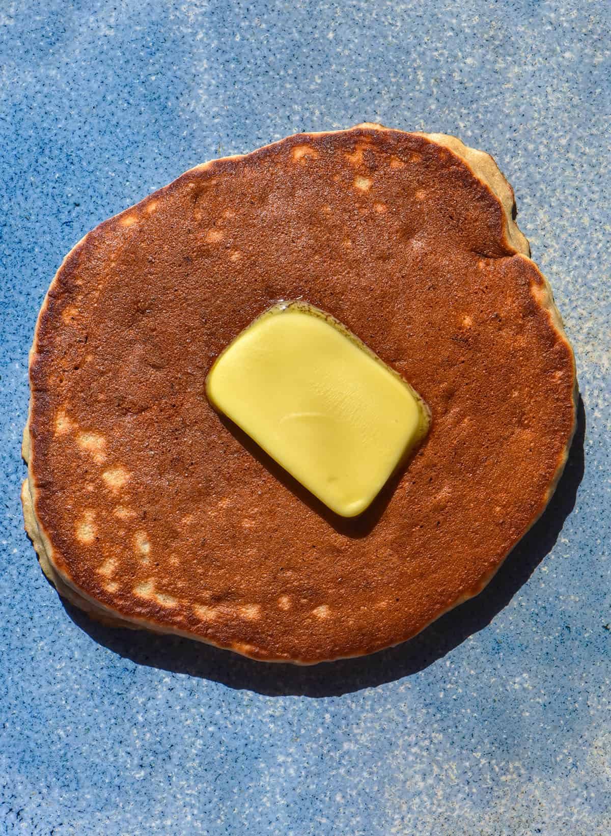 An aerial image of a buckwheat banana pancake on a bright blue ceramic plate. The pancake is a deep brown and topped with a square piece of melting butter.